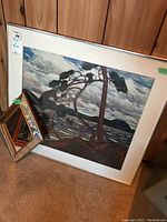 Framed print 'The West Wind' by Tom Thomson leaning against wood-paneled wall with small decorative mirror in front.