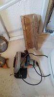 Full view of one Black & Decker circular saw without blade along with cardboard box for saw blade, cords visible on cement floor.