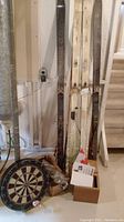 Three pairs of cross country skis, dart board with darts, some boxes, and parts of finishing net and pool cue beside wooden stairs.