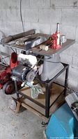 Table saw on metal stand with accessories like mallet, screwdriver, hydraulic jack and gloves in garage setting.