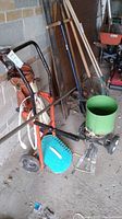 Photo showing multiple gardening and landscaping tools including rakes, shovel, hole digger, pipe, and crowbar leaning against wall.