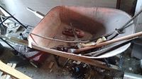 Metal wheelbarrow with wooden handles containing several tools inside including a sledgehammer, a trailer hitch part, and a come-along hand winch with chain. The wheelbarrow and tools show visible scratches and wear.