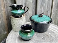 Three 1970s Blue Mountain Pottery pieces shown on a wooden surface outdoors with weathered fence background.