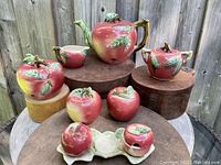 Photo showing entire 9-piece apple-themed ceramic tea set arranged on wooden blocks outdoors, featuring teapot, sugar bowl, salt and pepper shakers, large shakers, and jam bowl, all hand painted in red, yellow, green and brown colors with leaf and stem details.
