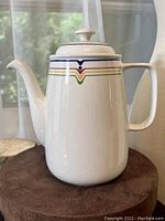 Front view of white ceramic coffee pot with colorful striped detail near the top and a handle on the right side.