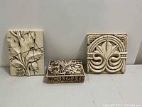 Three ivory tone carved resin wall hangings with botanical and geometric motifs arranged side by side