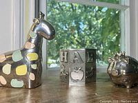 Photo of the silver plated giraffe bank with pastel spots, alphabet bank showing letters HAB, and princess pig bank with a crown, all on wooden surface by a window.