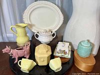 Overview of multiple pottery and china items on display including platter, covered sugar bowl, yellow pitcher, cow creamer, cups, turquoise jar, hexagonal jar, and decorative tile