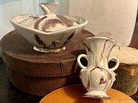 Photo showing the covered jar on a brown box beside a small vase with two handles, highlighting size and condition.