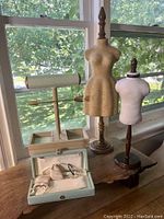 Three jewelry display items: two mannequin busts on wooden stands and a T-bar bracelet holder with compartments. Also visible is a light blue jewelry storage case open with cushions inside. Natural light from window background.