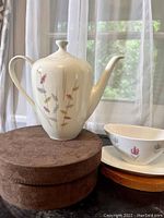 Front and side view of vintage white porcelain 6-cup teapot with slender multicolor floral decorations, placed on a brown round pedestal.