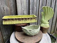 Three mid-century green ceramic items displayed outdoors on wooden disks against a wood fence: rectangular planter, swan vase, shell/conch planter