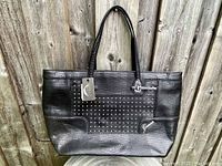 Front view of black Guess Rumi CarryAll tote with studded detail, logo, and hang tag visible, placed against a wood panel background.