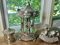 Overall view of the three silver plated carousel items together on wooden surface by window showing detailed designs including horses and floral motifs.