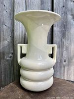 Front view of the cream Art Deco vase with flared rim and Greek key handles against a wooden fence background.