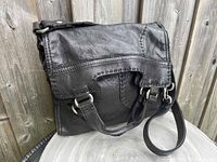 Front and side view of the black lamb leather handbag showing flap with rivet detailing, dual handles, adjustable shoulder strap attached with metal D-rings.