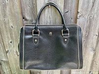 Front view of a black pebbled leather handbag with white trim on the edges, two top handles with metallic buckle attachments, and a small metallic Étienne Aigner logo at center front. The handbag is approximately 9x13 inches in size.