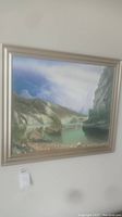 Framed mountain landscape painting featuring rocky mountains, greenery, and a water body under a sky, with a wide silver-colored frame.