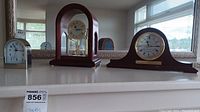 Shows the three clocks displayed on a white surface with a mirror background: small Linden desk clock on left, Seiko anniversary clock with wooden and glass case center, and Bulova mantel clock with curved wood on right.