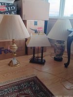 Three lamps positioned side by side on a wooden floor in front of boxes; includes brass lamp with pleated cream shade, wooden desk lamp with rectangular shade, and Asian style porcelain lamp with floral patterns.