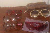Five pairs of sunglasses arranged on wooden surface with two cases. Various frame shapes and lens colors visible.
