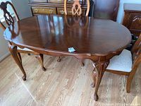 Wooden dining table with a polished dark finish, ornate carved legs, and scalloped edges. The top surface shows faint scuff marks. The table is extended with the leaf insert in place.