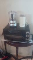 Photo showing DeLonghi toaster oven with Cuisinart Mini Prep Plus food processor and Mr. Coffee grinder on top of the toaster oven on a round table.