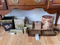 Photo showing a collection of toiletries accessories including multiple wastebaskets, soap holder, toothbrush holder, jar, tissue box, and three trays arranged on floor under wooden cabinet.