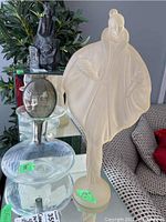 Image showing the Canadian Fashion Lady sculpture next to the blown glass decanter. Sculpture is frosted glass figure of a woman in a flowing cape. The decanter is clear glass with rounded body and spherical stopper.