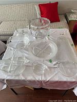 Multiple items of clear glass serveware arranged on white cloth, includes bowls, divided trays, and a cake stand with dome lid.