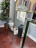 Full side view of mirrored stand showing reflective panels, standing on wooden floor next to decorative vases and plant