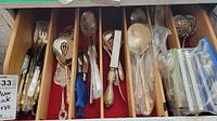 Top-down view of wooden tray compartments filled with assorted silver plate serveware and small spoons.