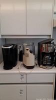 Three small kitchen appliances on countertop including a black Keurig coffee maker, a white Moulinex food processor, and a stainless steel Oster drip coffee maker with glass carafe. All plugged in but untested.