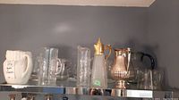 Wide view showing the entire lot on a glass shelf, including glass vases, clear glass pitchers, a white opaque vase, cut glass pitcher with gold lid, and silver plate pitcher with black handle.