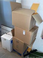 Four cardboard boxes stacked, two open showing yellow bubble mailers inside, whiteboard partially visible in background