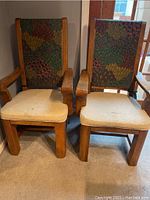 Pair of captain dining chairs with wooden frames, patterned black and multicolored fabric on backrests, and beige cushions showing wear and stains.