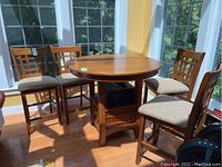 Four wood counter height chairs with upholstered beige seats and grid-back designs shown around a wood round table in sunlit room.