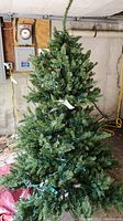6ft green artificial Christmas tree with dense branches pre-lit with multi-colored string lights standing on stand in garage environment with horseshoe and equipment in background. Red storage bag visible on floor.