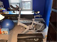 Side view of the stationary trainer and rolled black mat on carpeted floor under a window.