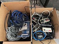 Box with tangled blue, gray, and black cords alongside black and gray Nintendo wired controllers with red buttons and attached cords.