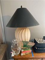 Full view of white ceramic lamp with large black shade, glass vase, stack of twist candles, and straw fan on a surface