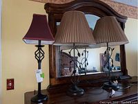 Three metal base table lamps. Two are matching with pleated tan shades and leaf accents; one is a single lamp with a red square shade.