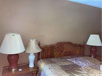 Three ceramic table lamps on bedside furniture; two reddish matching lamps with pleated shades and one white lamp with damaged shade in the center.