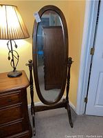 Full view of the oval floor mirror with wooden frame and stand placed near furniture and lamp.
