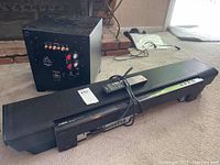 Photo showing Martin Logan Dynamo subwoofer and Yamaha YSP-4000 sound bar with remote on carpeted floor near a fireplace.