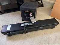 Image showing the Yamaha YSP-1100 sound bar lying on floor with remote control on top and Martin Logan Dynamo subwoofer behind.