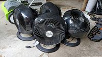 Four black Vornado fans grouped together on a surface. All fans have the Vornado logo visible on the front grills. Fans appear to be from the same model line with round bases and upward tilting fan heads.