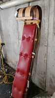 Full view of the wooden toboggan leaning against a wall, showing the red padded seat and yellow ropes.