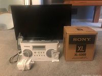 Photo showing Samsung TV behind Sandian stereo receiver, Garmin pager, and Sony XL-5300 lamp unit box.