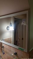 Large rectangular bathroom mirror with cream/off-white wooden frame mounted above beige tile backsplash and next to a light fixture. Shows wear and is in a bathroom setting.